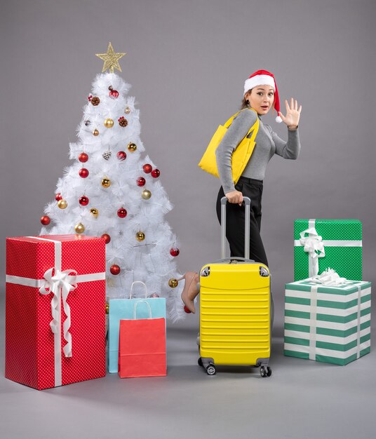 Mujer vistiendo gorro de Papá Noel con equipaje junto al árbol de Navidad