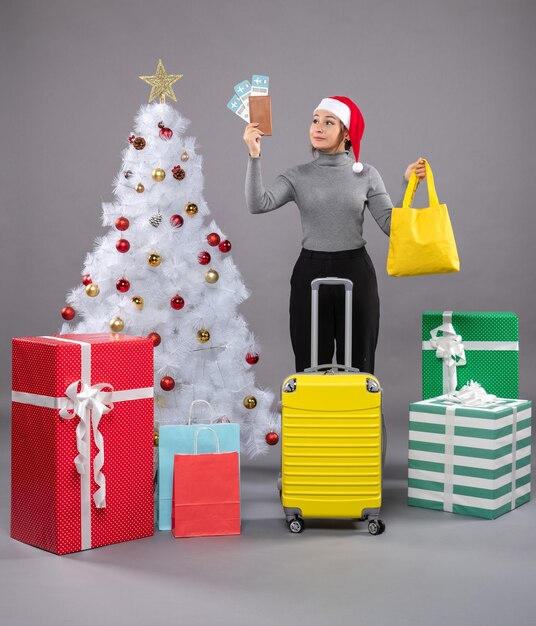 Mujer vistiendo gorro de Papá Noel con equipaje junto al árbol de Navidad