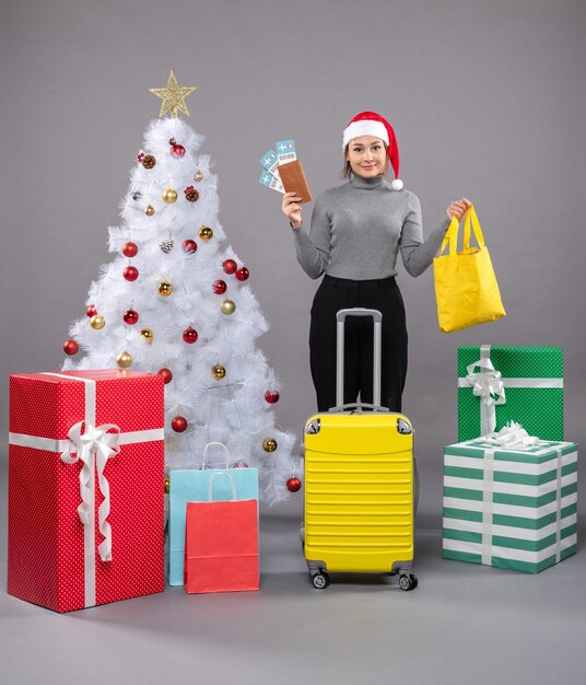 Mujer vistiendo gorro de Papá Noel con equipaje junto al árbol de Navidad