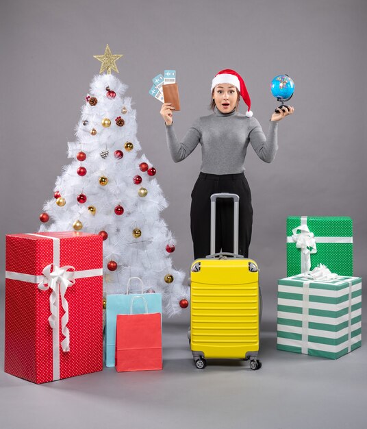 Mujer vistiendo gorro de Papá Noel con equipaje junto al árbol de Navidad