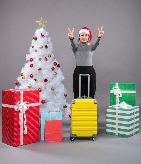 Mujer vistiendo gorro de Papá Noel con equipaje junto al árbol de Navidad