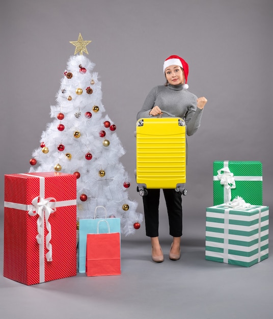 Mujer vistiendo gorro de Papá Noel con equipaje junto al árbol de Navidad