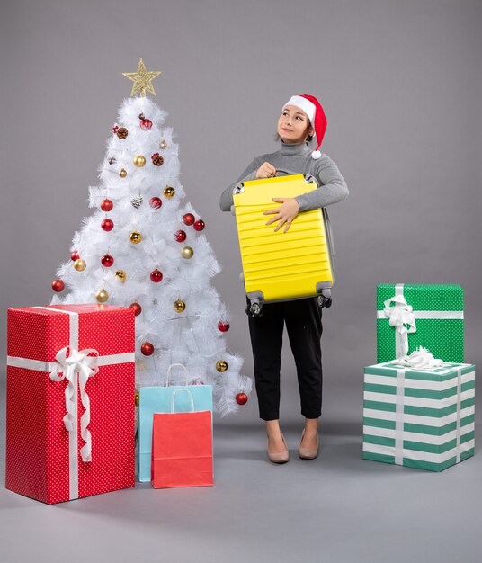 Mujer vistiendo gorro de Papá Noel con equipaje junto al árbol de Navidad