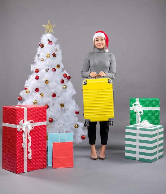 Mujer vistiendo gorro de Papá Noel con equipaje junto al árbol de Navidad