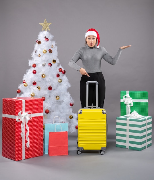 Mujer vistiendo gorro de Papá Noel con equipaje junto al árbol de Navidad