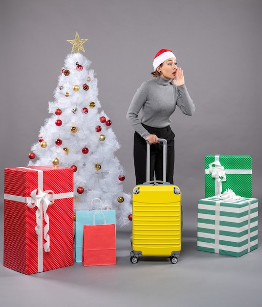 Mujer vistiendo gorro de Papá Noel con equipaje junto al árbol de Navidad