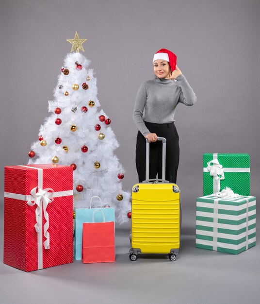 Mujer vistiendo gorro de Papá Noel con equipaje junto al árbol de Navidad