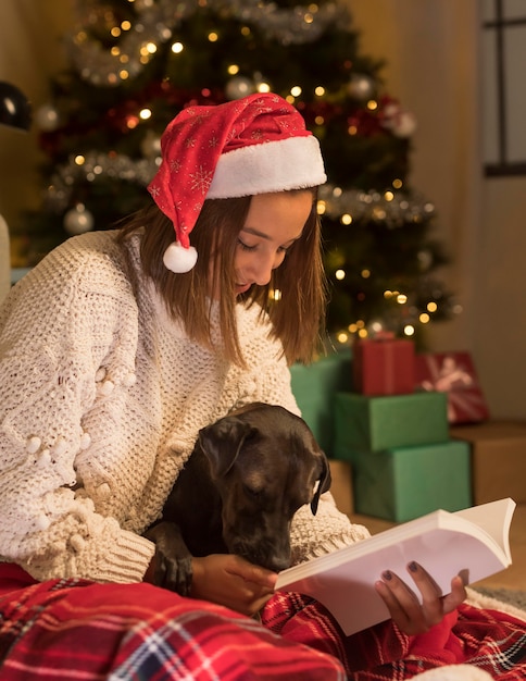 Mujer vistiendo gorro de Papá Noel en Navidad y libro de lectura con su perro