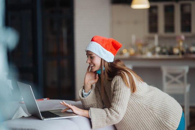 Mujer vistiendo santa sonriendo mientras habla con un amigo en línea en la computadora portátil durante la celebración de Navidad en casa