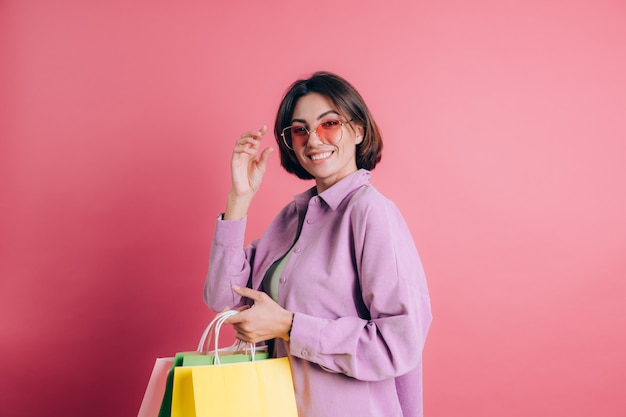 Mujer vistiendo un suéter casual sobre fondo feliz disfrutando de compras sosteniendo bolsas coloridas con gafas de sol de verano