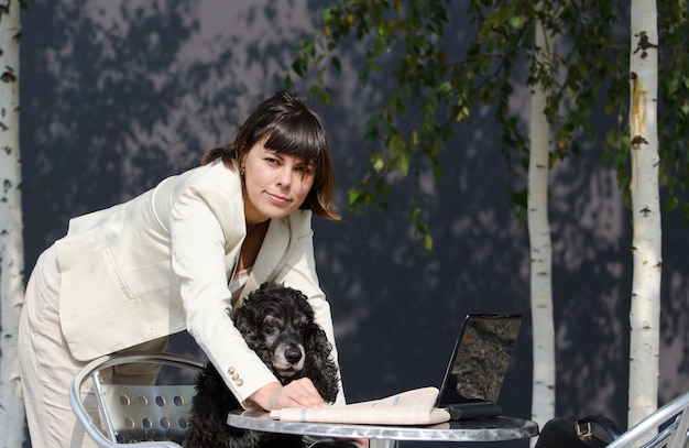 Mujer vistiendo un traje blanco sosteniendo a su perro y usando su computadora portátil sobre la mesa