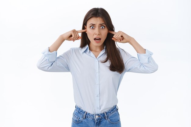 Mujer volviéndose loca por la música fuerte y horrible que toca a sus vecinos, quejándose de ruido haciendo muecas molestas y disgustadas, siente una gran incomodidad enchufar los dedos en los oídos, pared blanca