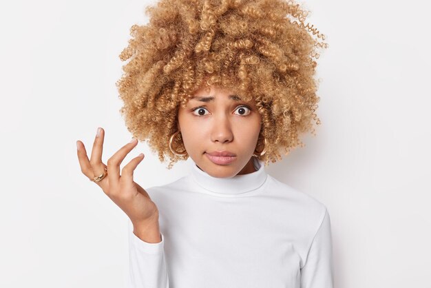 La mujer ypung de cabello rizado, perpleja y vacilante, se encoge de hombros con desconcierto, se siente confundida, usa un puente casual aislado sobre fondo blanco. Entonces que hacer en esta dificil situacion me pregunto