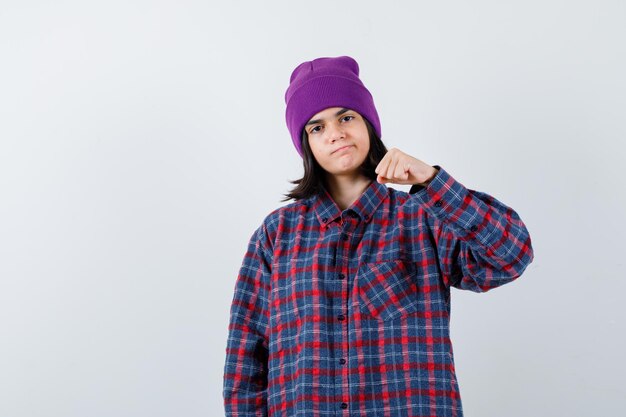 Mujercita en camisa a cuadros y gorro levantando puño cerrado mirando confiado