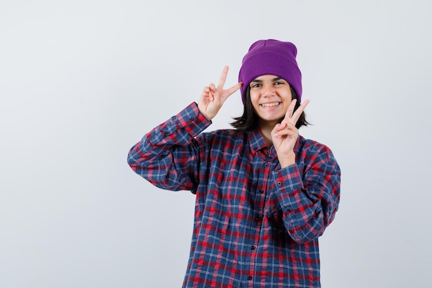 Mujercita mostrando el signo de la victoria en camisa a cuadros y gorro y mirando alegre