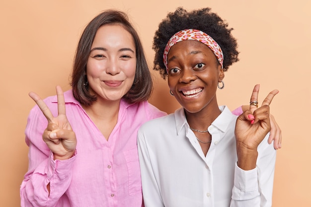 Las mujeres se abrazan y muestran el gesto de la victoria, la sonrisa demuestra positivamente la paz, el signo de la discoteca se divierten juntos vestidos casualmente aislados en beige