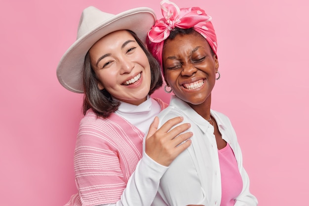 las mujeres se abrazan y se paran cerca una de la otra sonríen ampliamente usan ropa elegante aislada en rosa