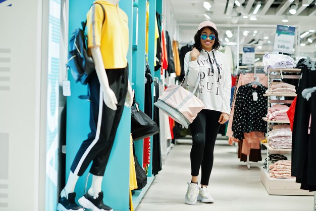 Mujeres afroamericanas en chándales y gafas de sol de compras en el centro comercial de ropa deportiva con bolsa deportiva contra estantes Tema de la tienda deportiva