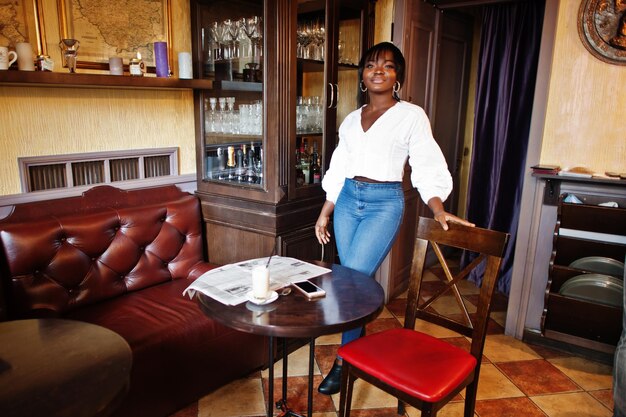 Mujeres afroamericanas con estilo en blusa blanca y jeans azules posaron en el café con periódico