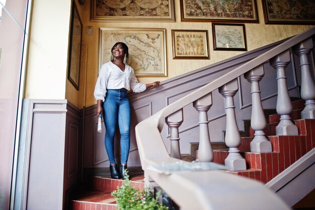 Mujeres afroamericanas con estilo en blusa blanca y jeans azules posaron en el café con periódico