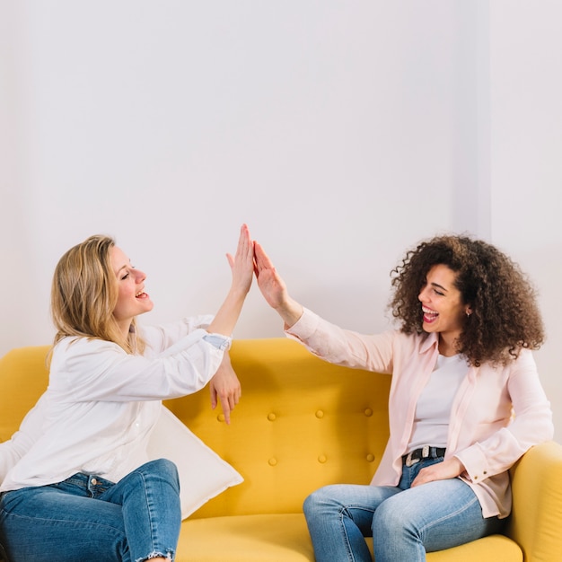 Mujeres alegres alto-fiving en el sofá amarillo