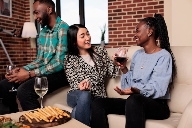 Mujeres alegres multiétnicas en la fiesta del vino hablando mientras se sientan en el sofá. Felices y sonrientes amigos diversos sentados en la sala de estar mientras disfrutan de bebidas alcohólicas juntos mientras celebran el evento de cumpleaños.