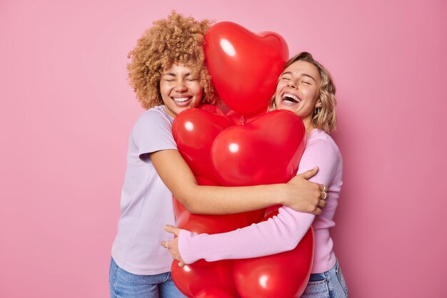Las mujeres alegres y optimistas abrazan un gran montón de globos inflados en forma de corazón vestidos con ropa informal y se divierten preparándose para la celebración del cumpleaños o el día de San Valentín aislado sobre fondo rosa