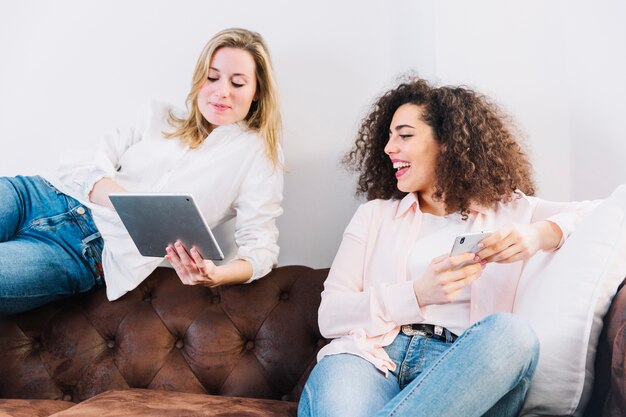 Mujeres alegres que usan tecnologías en el sofá