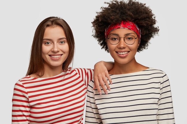 Mujeres alegres satisfechas de diferentes razas, sentirse felices con una charla divertida, sonreír ampliamente, usar suéteres a rayas similares, aislados sobre una pared blanca, tener un gran humor y diversión. Chicas multiculturales