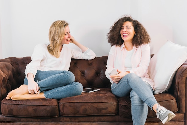 Mujeres alegres con tecnologías hablando en sofá