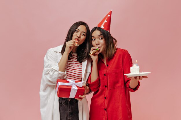 Mujeres asiáticas bronceadas soplan cuernos de fiesta en aislado Linda mujer en blusa roja y sombrero de fiesta sostiene pastel de cumpleaños Jovencita en traje elegante posa con caja de regalo sobre fondo rosa