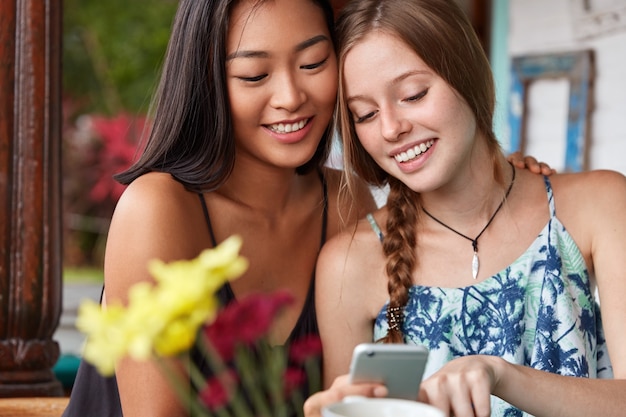 Las mujeres asiáticas y caucásicas alegres positivas tienen expresiones alegres, pasan tiempo juntas, miran videos en blogs en teléfonos inteligentes