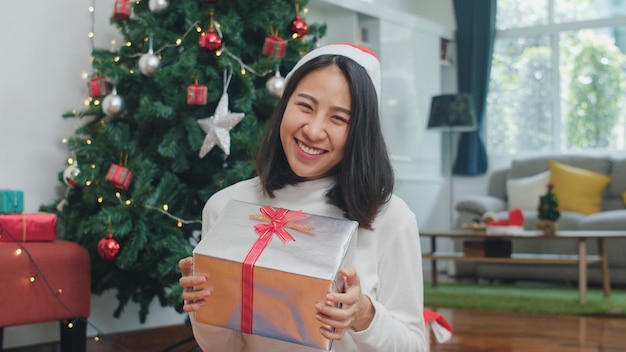 Las mujeres asiáticas celebran el festival de Navidad. Adolescente femenino use suéter y sombrero de Navidad relajarse feliz espera regalo sonriendo cerca del árbol de Navidad disfrutar de vacaciones de invierno de Navidad juntos en la sala de estar en casa