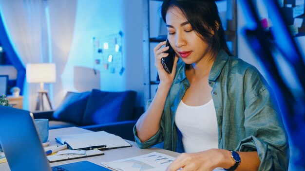 Mujeres asiáticas independientes que usan la computadora portátil hablan por teléfono a un empresario ocupado que trabaja a distancia en la sala de estar.