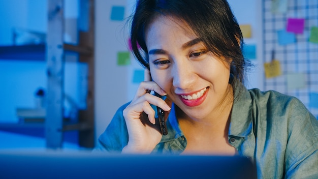 Mujeres asiáticas independientes que usan la computadora portátil hablan por teléfono a un empresario ocupado que trabaja a distancia en la sala de estar.