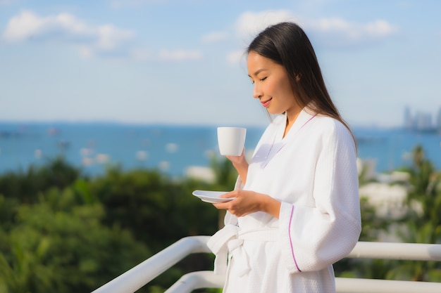 Las mujeres asiáticas jovenes hermosas del retrato sostienen la taza de café en mano alrededor de la visión al aire libre