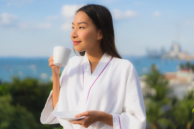 Las mujeres asiáticas jovenes hermosas del retrato sostienen la taza de café en mano alrededor de la visión al aire libre