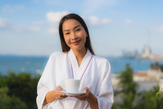 Las mujeres asiáticas jovenes hermosas del retrato sostienen la taza de café en mano alrededor de la visión al aire libre