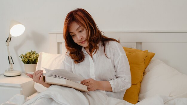 Las mujeres asiáticas mayores se relajan en casa. La hembra china mayor asiática disfruta del libro de lectura del tiempo de descanso mientras que miente en cama en dormitorio en casa en el concepto de la noche.