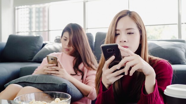 Mujeres asiáticas que usan teléfonos inteligentes y comen palomitas de maíz en la sala de estar en casa, grupo de compañeros de habitación amigos