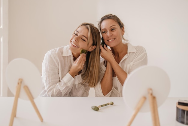 Foto gratuita mujeres bastante caucásicas con raspador de gouache de jade, rubias haciendo masajes faciales mientras se miran en el espejo. belleza y cuidado personal, concepto de cuidado de la piel.