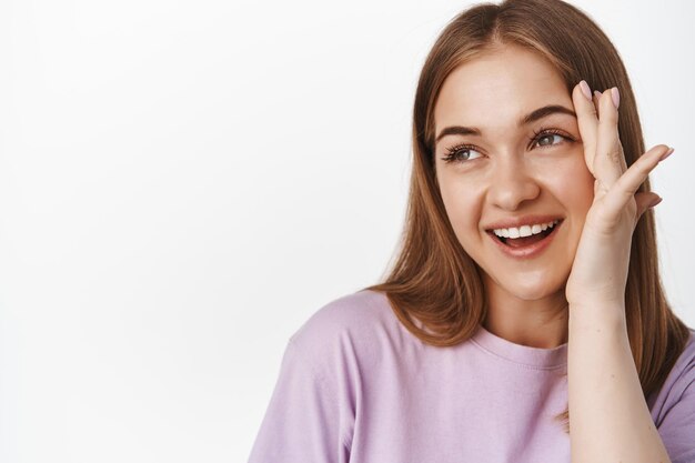 Mujeres de belleza. Cierra el retrato de la cara de una mujer hermosa, la chica toca su piel facial y sonríe, sonrojándose y mirando el espacio para texto promocional, fondo blanco.