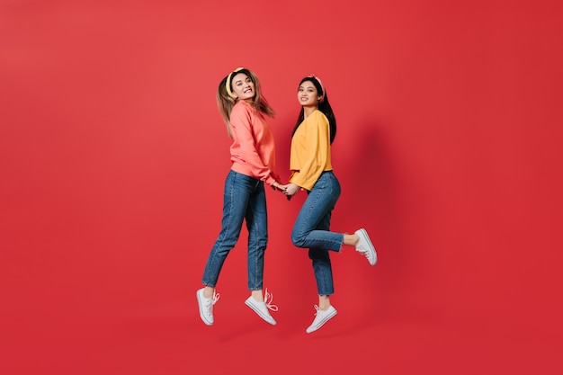 Mujeres bonitas en elegantes sudaderas y jeans saltan sobre la pared roja