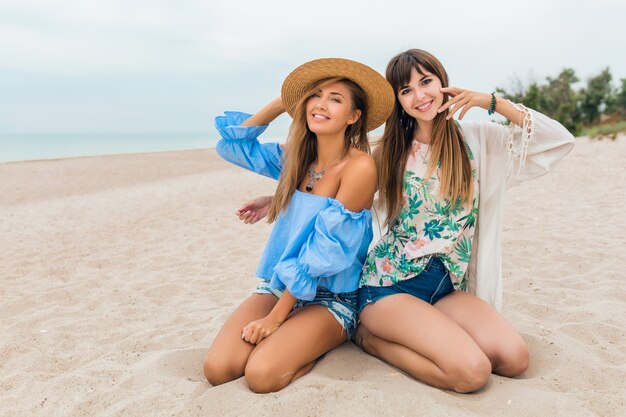 Mujeres bonitas con estilo sentadas en la arena en las vacaciones de verano en la playa tropical, estilo bohemio, amigos viajan juntos, accesorios de moda, emoción feliz sonriente, estado de ánimo positivo, sombrero de paja