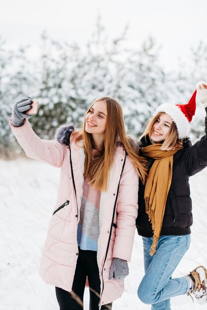 Mujeres bonitas en el sombrero de Papá Noel que toma el selfie en bosque del invierno