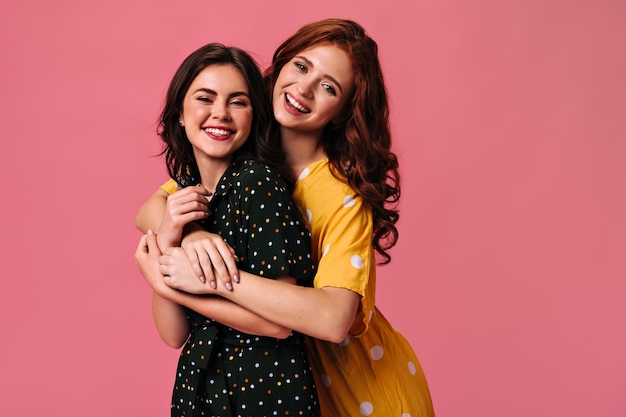 Foto gratuita mujeres con cabello ondulado abrazándose sobre fondo rosa chicas maravillosas con ropa hermosa y colorida y con labios brillantes linda sonrisa a la cámara