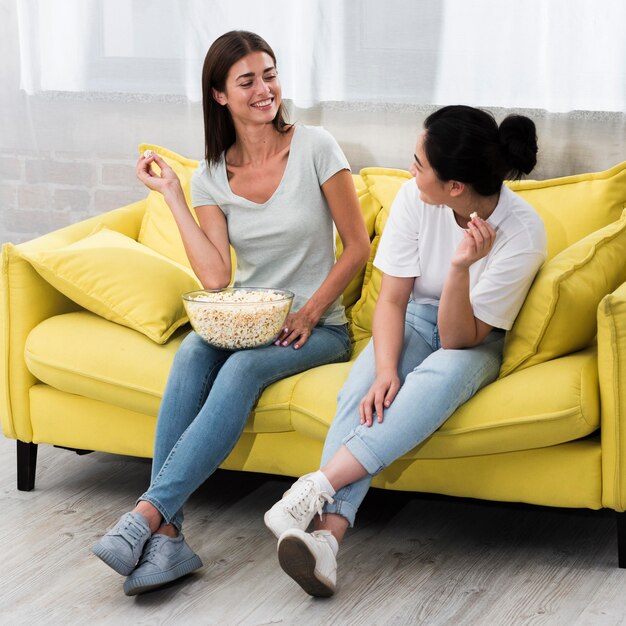 Mujeres en casa en el sofá charlando y comiendo palomitas de maíz