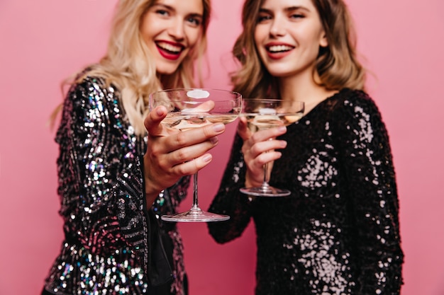 Mujeres caucásicas felices en vestidos negros posando en la fiesta. Chicas complacidas en traje brillante bebiendo vino juntas.