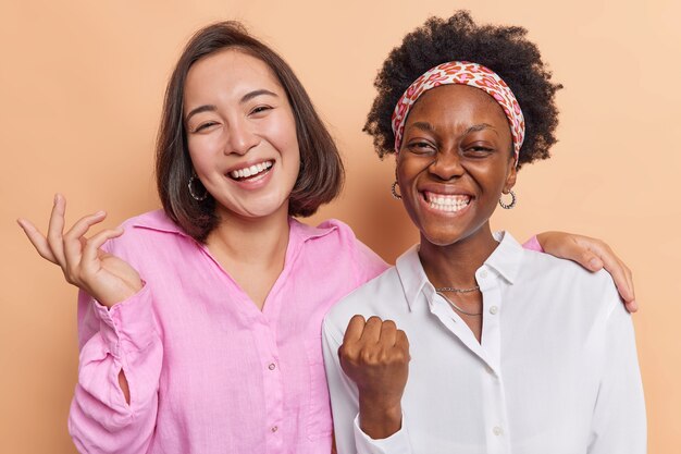 las mujeres celebran los logros se sienten muy positivas sonríen ampliamente de pie cerca unas de otras vestidas con camisas en beige