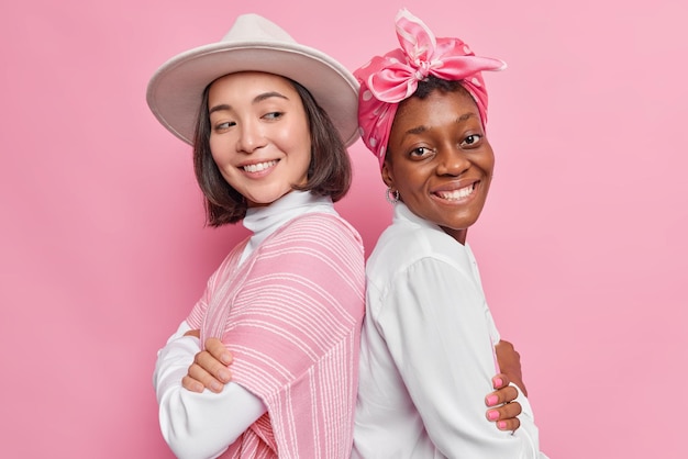 las mujeres se colocan de espaldas, mantienen los brazos cruzados, sonríen con gusto, trabajan como equipo aislado en rosa. Alegre modelo de mujer de piel oscura pasa tiempo libre con su hermana.